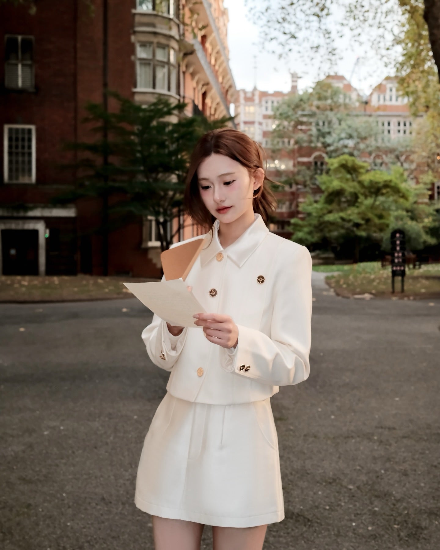 一位身穿 the.lookbook.select 白色 Grace Little Jacket 的女士站在戶外的鋪面上閱讀紙張，背景是樹木和磚造建築物。.