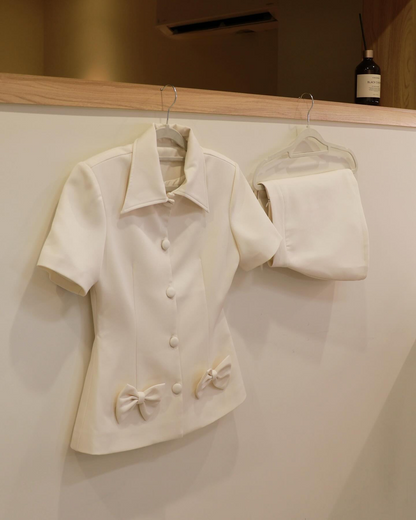 Light Skirt Set 🖤新品優惠 2件9折，由 lookbook.select 設計，包括一件帶蝴蝶結飾邊的白色短袖鈕扣上衣和一件同款短裙，掛在衣架上，靠著一面淺色的牆。.