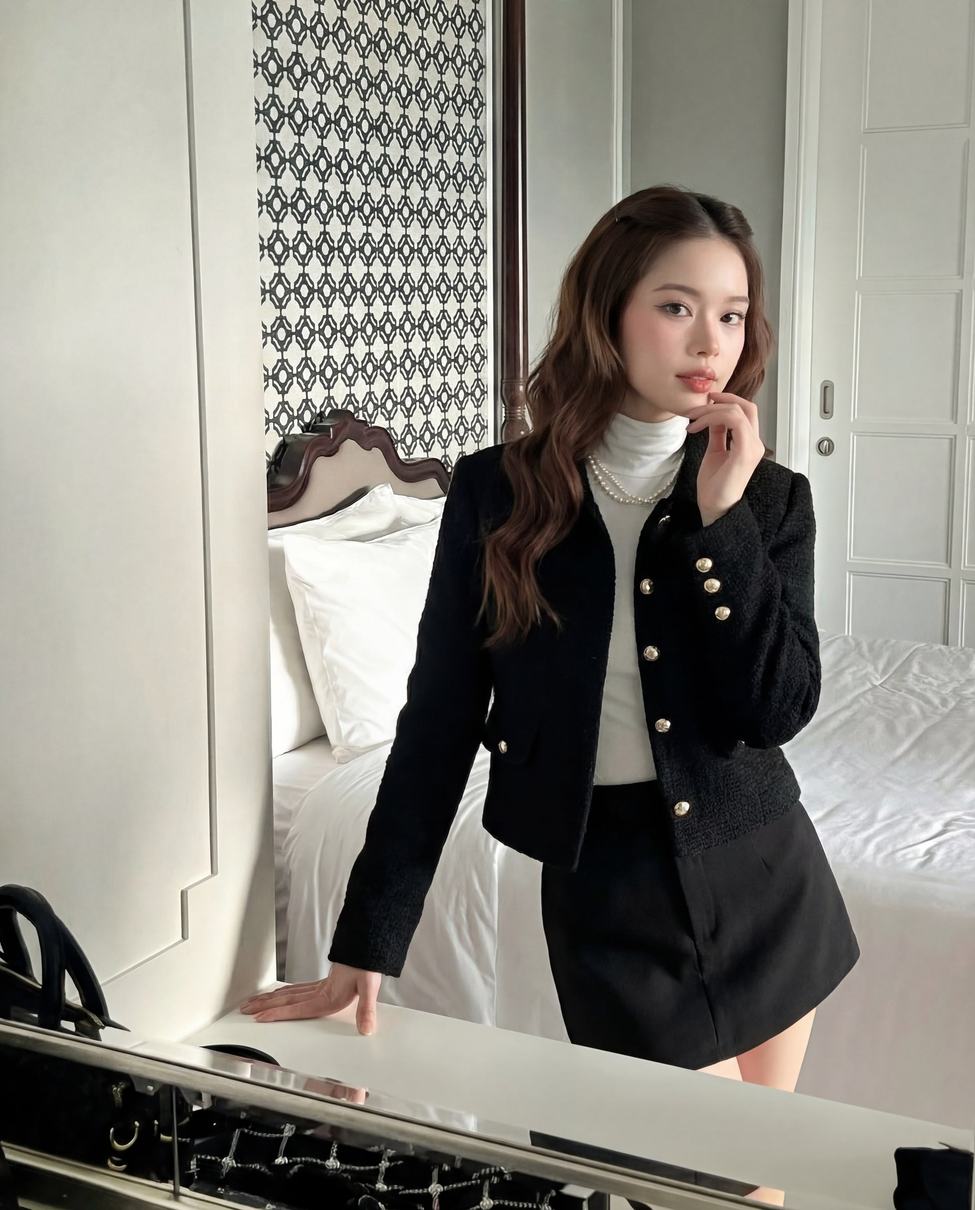 一位身穿 the.lookbook.select 的學院粗花呢外套和裙子的女士站在現代臥室的床邊，一隻手放在白色書桌上。.