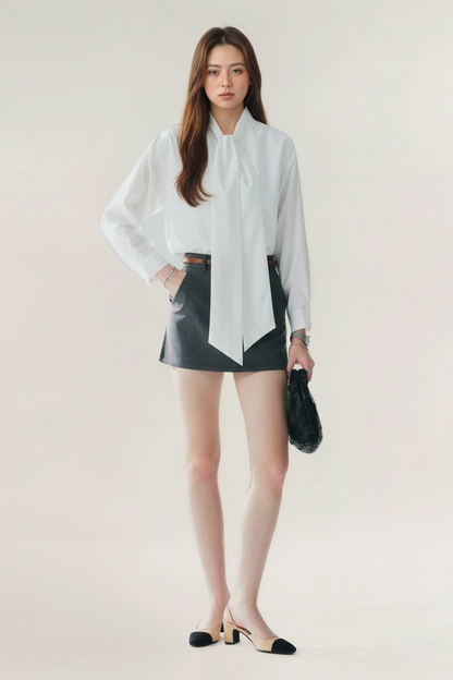 一位女士在素色背景下擺姿勢，穿著 the.lookbook.select 的蝴蝶結寬鬆襯衫，搭配黑色迷你裙、雙色高跟鞋和黑色手袋。.