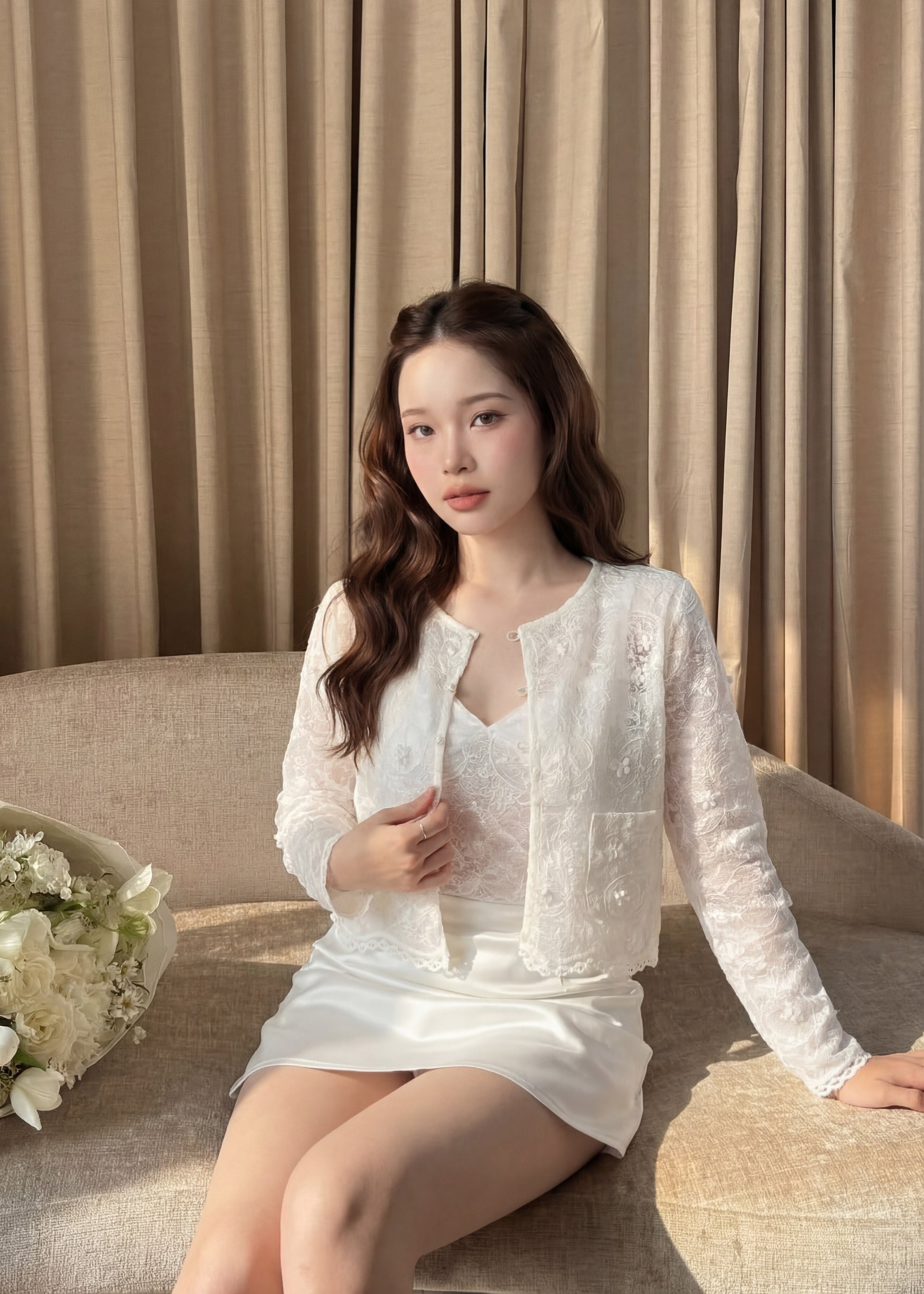 一位女士穿著 the.lookbook.select 的 Lumiere Lace Duo (Cami & Jacket) 坐在窗簾前的米色沙發上，身旁有一束白色鮮花。.