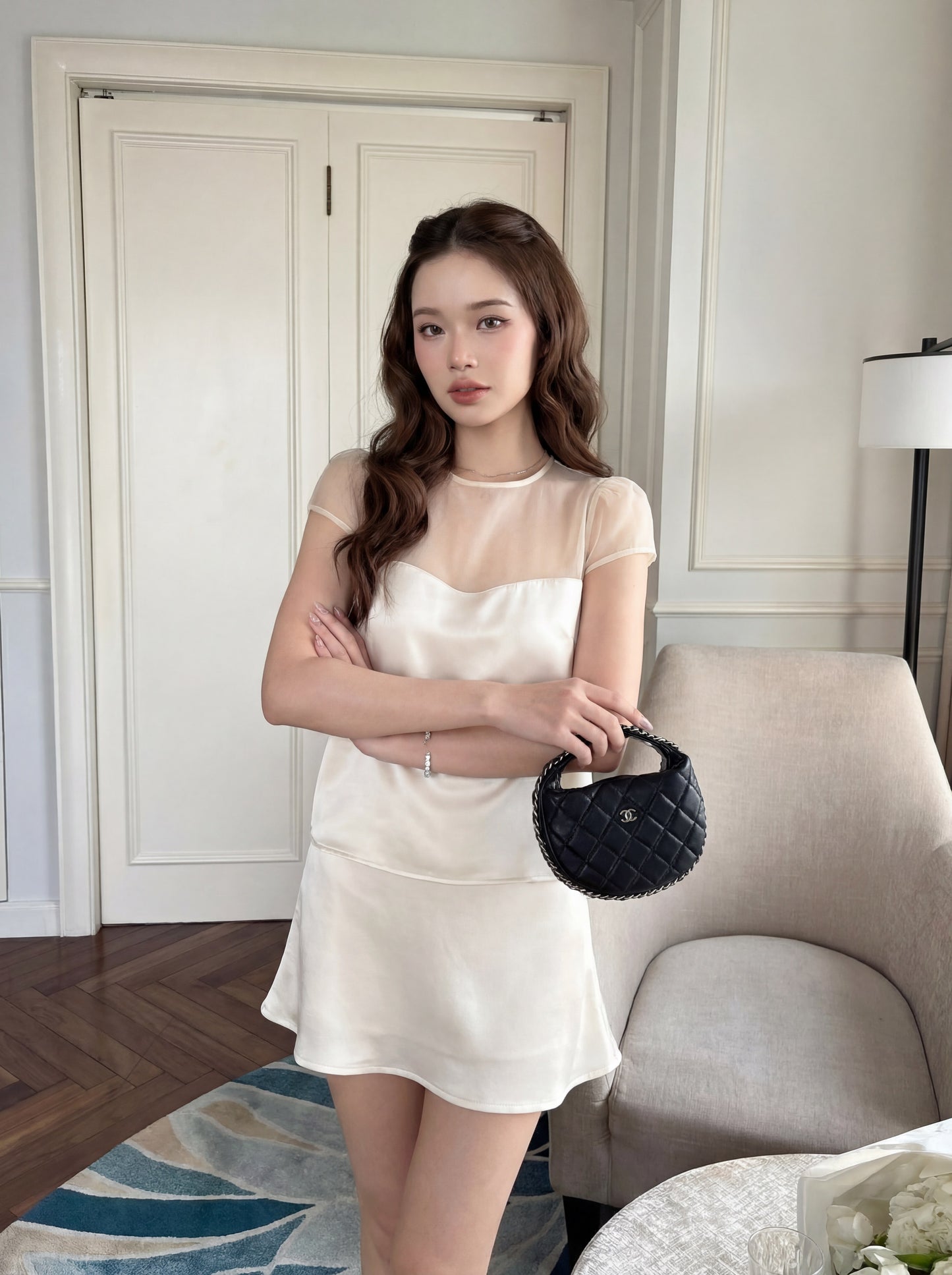 一名身穿.lookbook.select「緞面夢幻套裝」（上衣與裙子）的女子，雙臂交叉，站在室內，手拿一個小巧的黑色手提包，身旁是一張米色扶手椅和一張白色桌子。.