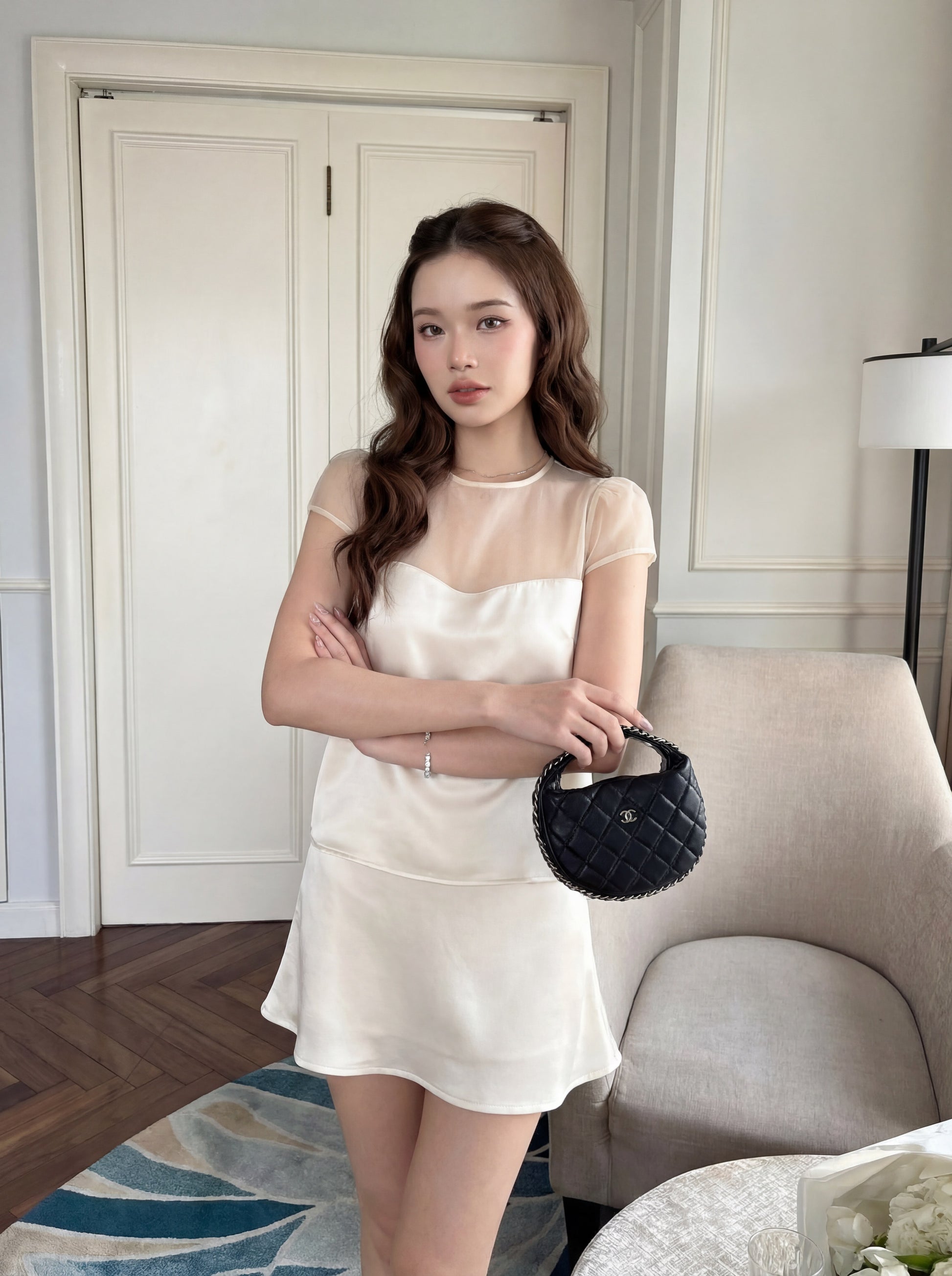 一名身穿.lookbook.select「緞面夢幻套裝」（上衣與裙子）的女子，雙臂交叉，站在室內，手拿一個小巧的黑色手提包，身旁是一張米色扶手椅和一張白色桌子。.