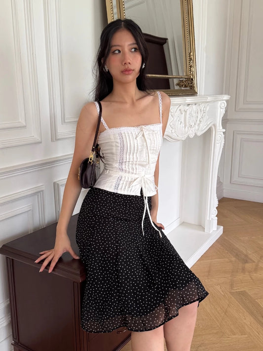 一位身穿白色無袖上衣和the.lookbook.select's Pois Midi Skirt的女士坐在一個優雅的白色房間的木桌上，房間裡有一面鏡子和華麗的壁爐，展現法式風情。.