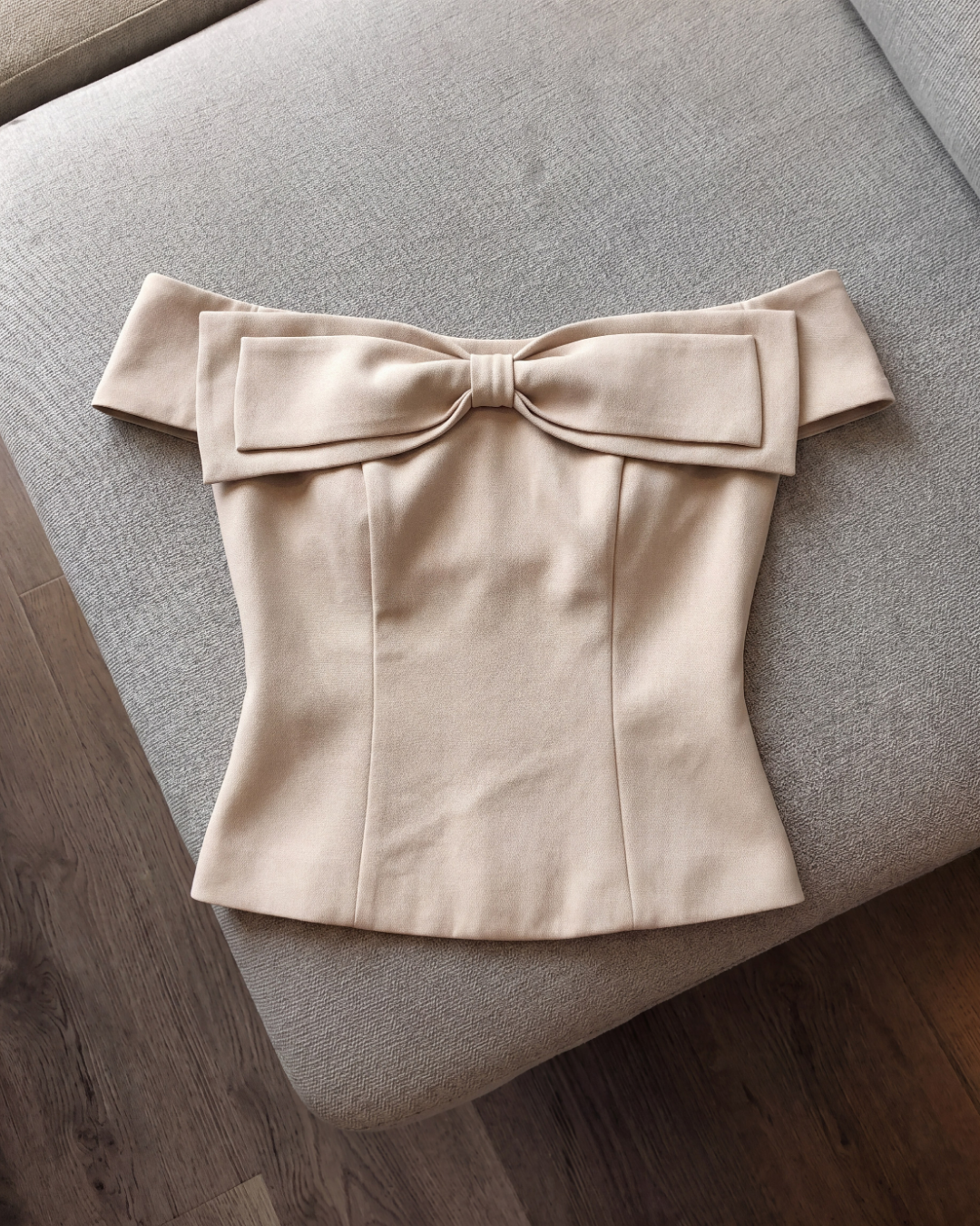 這款由 the.lookbook.select 推出的 Aria Bow Top 是一件米色法式一字領蝴蝶結上衣，胸前飾有大型蝴蝶結，並採用修身剪裁，圖中展示於淺灰色軟墊表面上。.