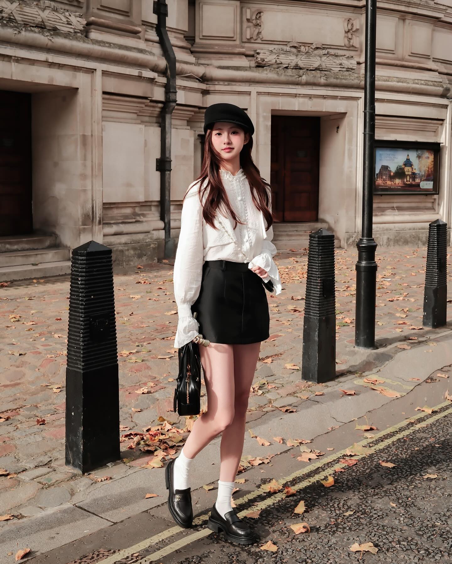 一位穿著 Hazel White Shirt 的法式氣質女性站在大樓旁，造型來自 the.lookbook.select，搭配黑色短裙、帽子、鞋子、白色襪子和手袋。落葉散落在人行道上。🖤新品優惠至12月5日.
