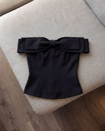 這款由 the.lookbook.select 推出的「Aria Bow Top」，是一件配有大型蝴蝶結且採用修身剪裁的黑色蝴蝶結上衣，在木質地板旁的一張米色沙發上展示，突顯其法式一字領的優雅風采。.