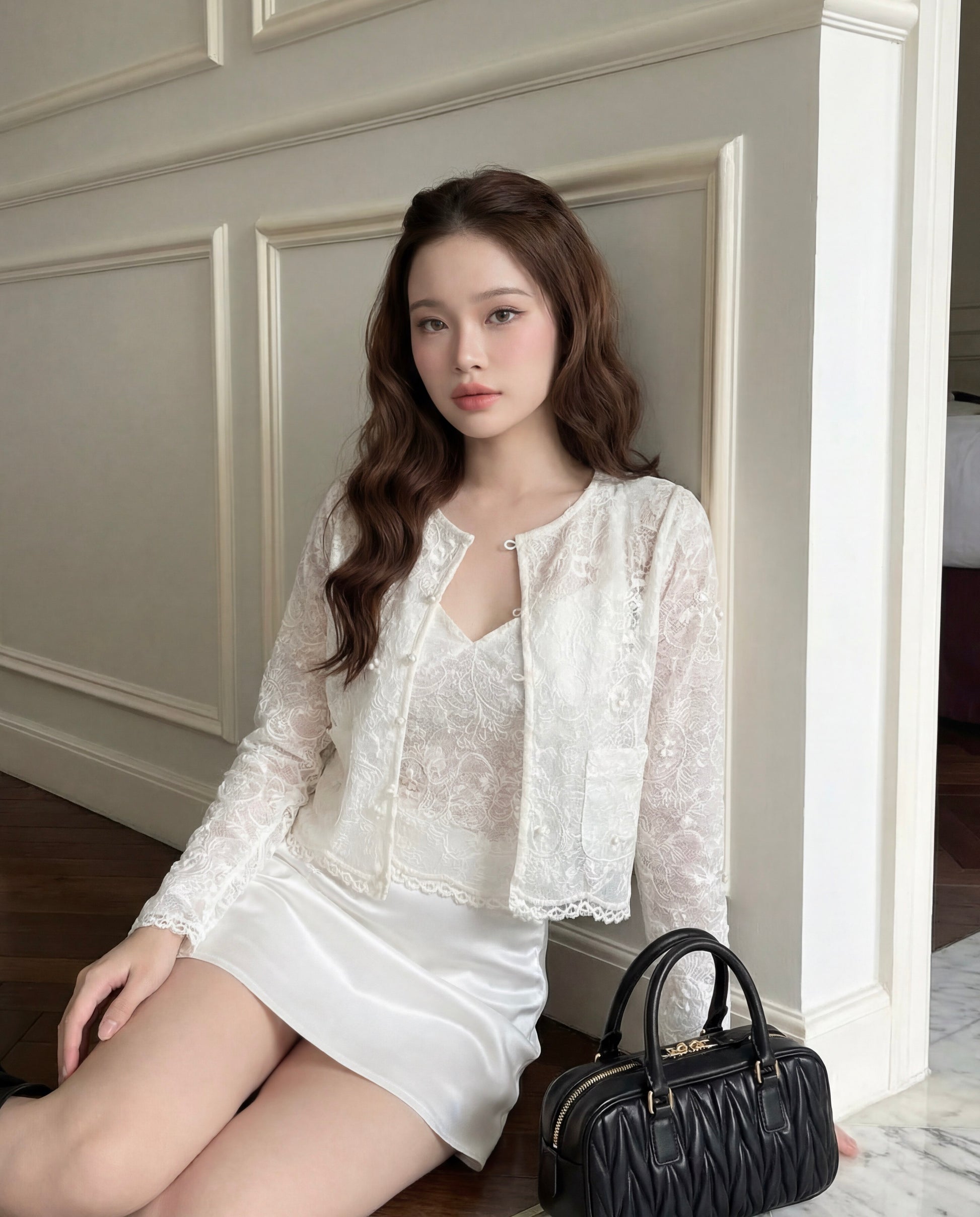 一位留著長棕髮的女子坐在鋪有木板牆的地板上，身穿 the.lookbook.select 的「Lumiere Lace Duo」（吊帶背心與外套）米白色套裝，身旁放著一個黑色手提包。.