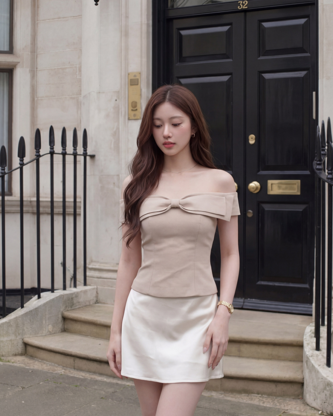 一名女子身穿 the.lookbook.select 品牌的米色 Aria Bow Top，這款上衣採用法式露肩設計且版型修身，她將其搭配白色裙子，佇立在黑色 32 號門前，身旁是石製欄杆。.
