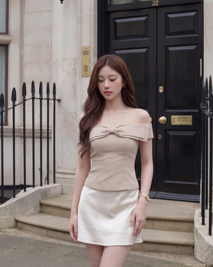 一名女子身穿 the.lookbook.select 品牌的米色 Aria Bow Top，這款上衣採用法式露肩設計且版型修身，她將其搭配白色裙子，佇立在黑色 32 號門前，身旁是石製欄杆。.