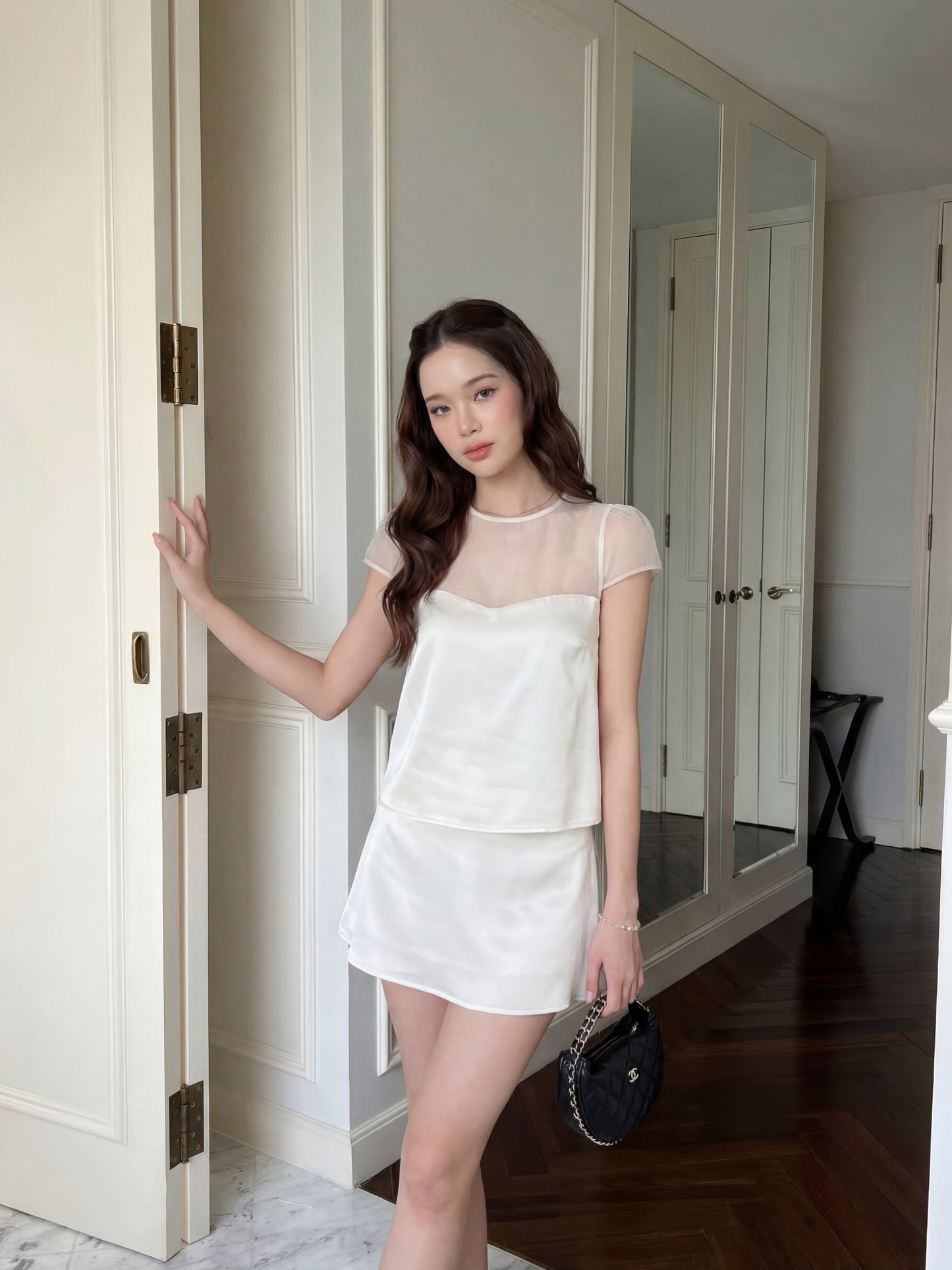 一名女子站在室內門邊，身穿 the.lookbook.select 的「Satin Reverie」套裝（上衣與裙子），左手拿著一個小巧的黑色手提包。.