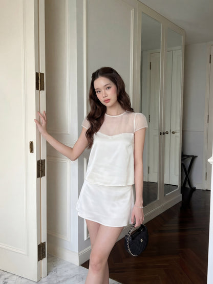 一名女子站在室內門邊，身穿 the.lookbook.select 的「Satin Reverie」套裝（上衣與裙子），左手拿著一個小巧的黑色手提包。.