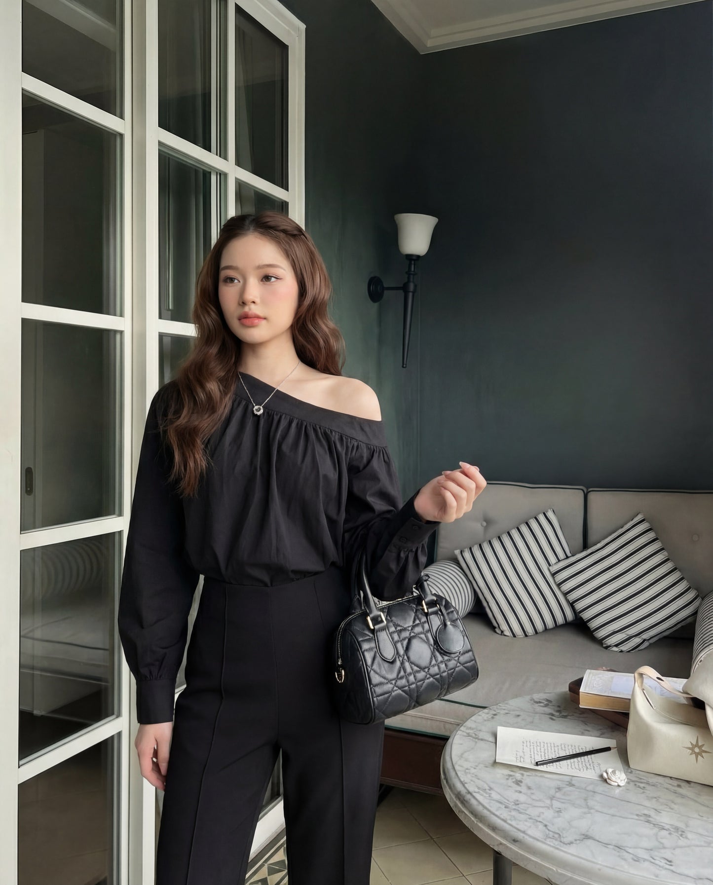一位身穿 The.lookbook.select 的 Lou Drape 襯衫和黑色褲子的女士站在室內，手拿黑色手提包，旁邊是一張放著書籍和飲料的桌子，這個現代化的休閒區有條紋靠墊。.