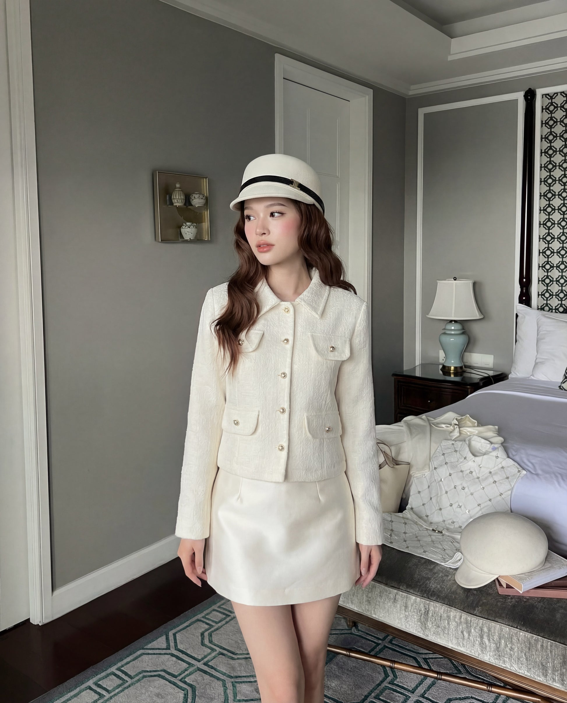 一位女士身著乳白色 College Tweed Coat by the.lookbook.select，搭配短裙，戴著白色帽子，站在中性裝潢、床舖整齊的現代臥室中。.