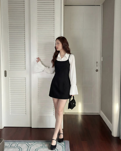 一位女士站在室內，身穿 the.lookbook.select 的 Muse Cami 連身裙，纖細肩帶和合身剪裁，搭配喇叭袖白色上衣。她拿著一個黑色小手包，凝視著旁邊。.
