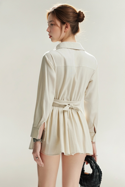 一位梳著髮髻的女士面對著遠方，她穿著 the.lookbook.select 的 Moiselle 襯衫連衣裙 - 一件輕盈的長袖束帶連衣裙，同時拿著一個黑色編織手提包。.