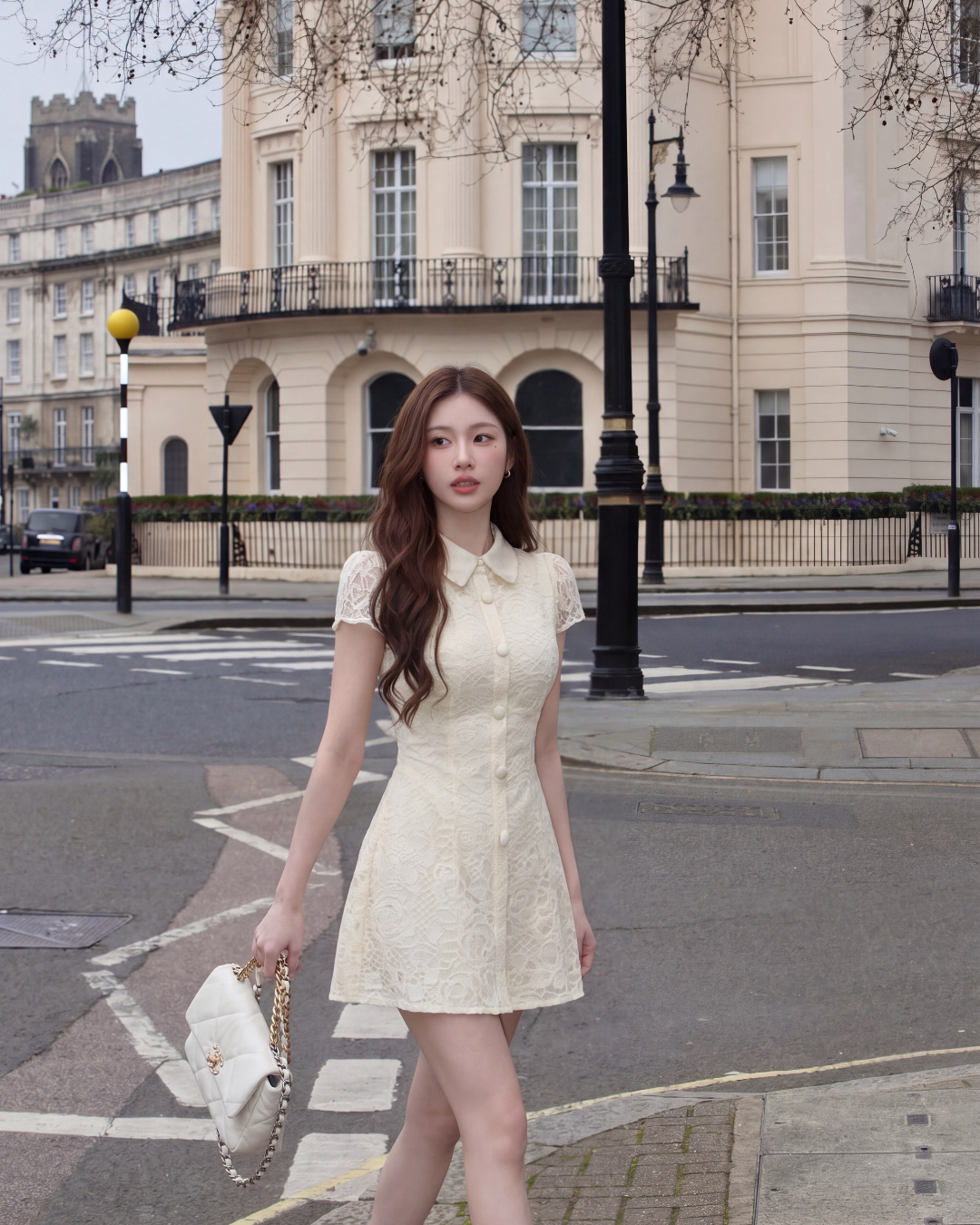 一位身穿 the.lookbook.select 惠靈頓蕾絲連身裙的女士，手持白色棉被手提包，走在有高大乳白色建築物和樹木的城市街道上。.