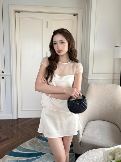 一位身穿.lookbook.select「緞面夢幻套裝」（上衣與裙子）的女性，手持一個小巧的黑色手提包，站在室內，在光線充足的現代風格房間裡，以一張米色椅子為背景擺出姿勢。.