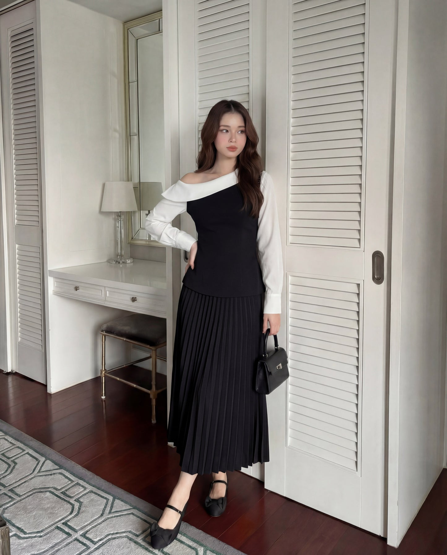 在室內，一位女士穿著 the.lookbook.select 的 Mono Skirt Set，搭配黑色不對稱上衣和百褶裙，手持黑色小手包。長凳、台燈和鏡子讓這個經典造型更為完整。.