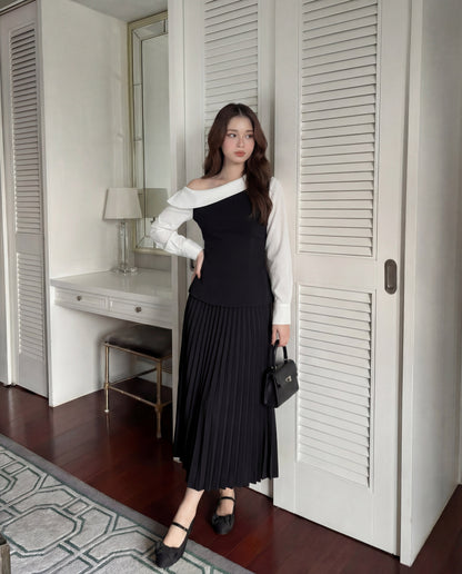 在室內，一位女士穿著 the.lookbook.select 的 Mono Skirt Set，搭配黑色不對稱上衣和百褶裙，手持黑色小手包。長凳、台燈和鏡子讓這個經典造型更為完整。.