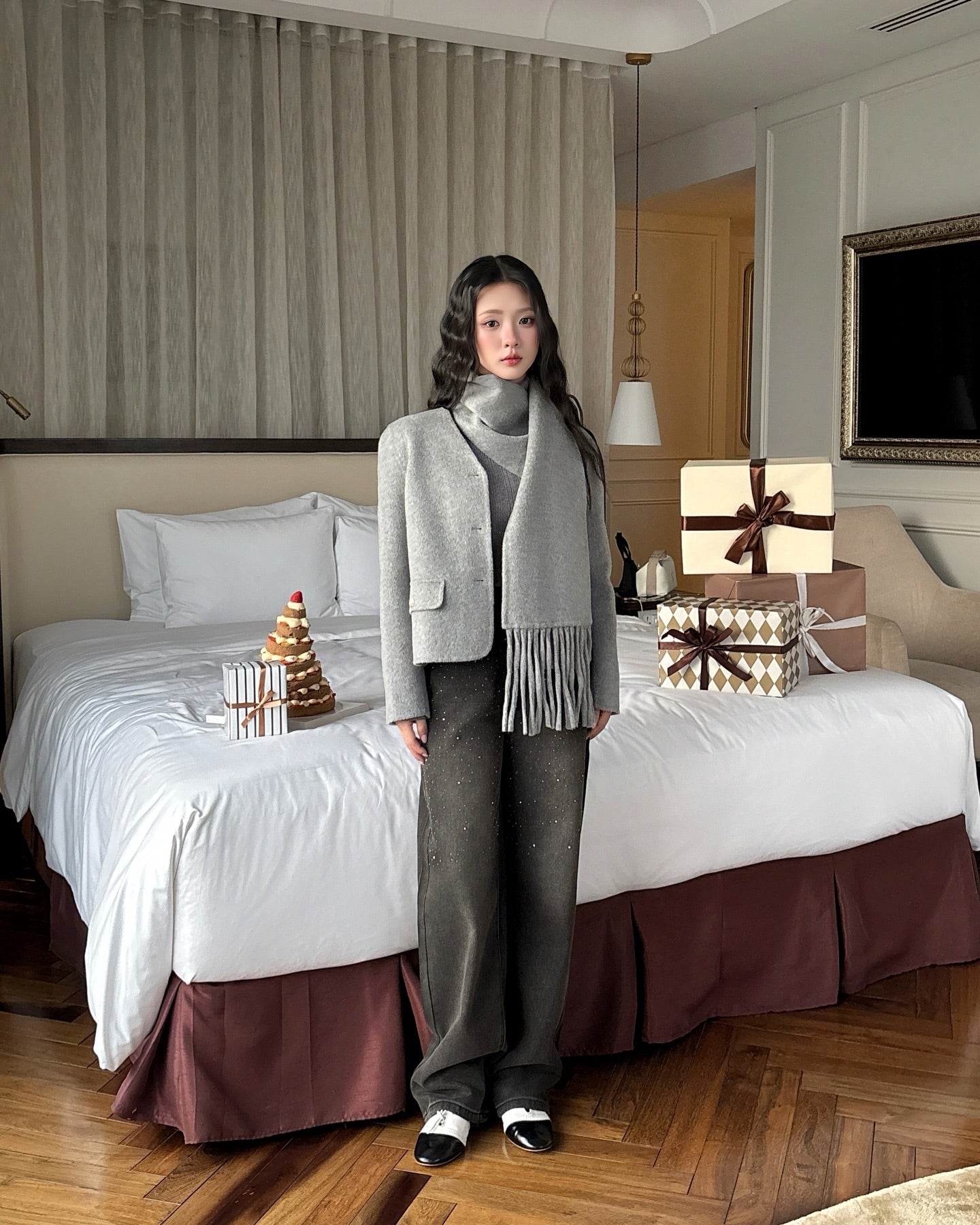 一位女士站在飯店房間裡，身穿 the.lookbook.select's Snow Wool Coat❄️、灰色流蘇圍巾和深色褲子。她身後的床上放著禮物盒和一棵裝飾用的小樹。.
