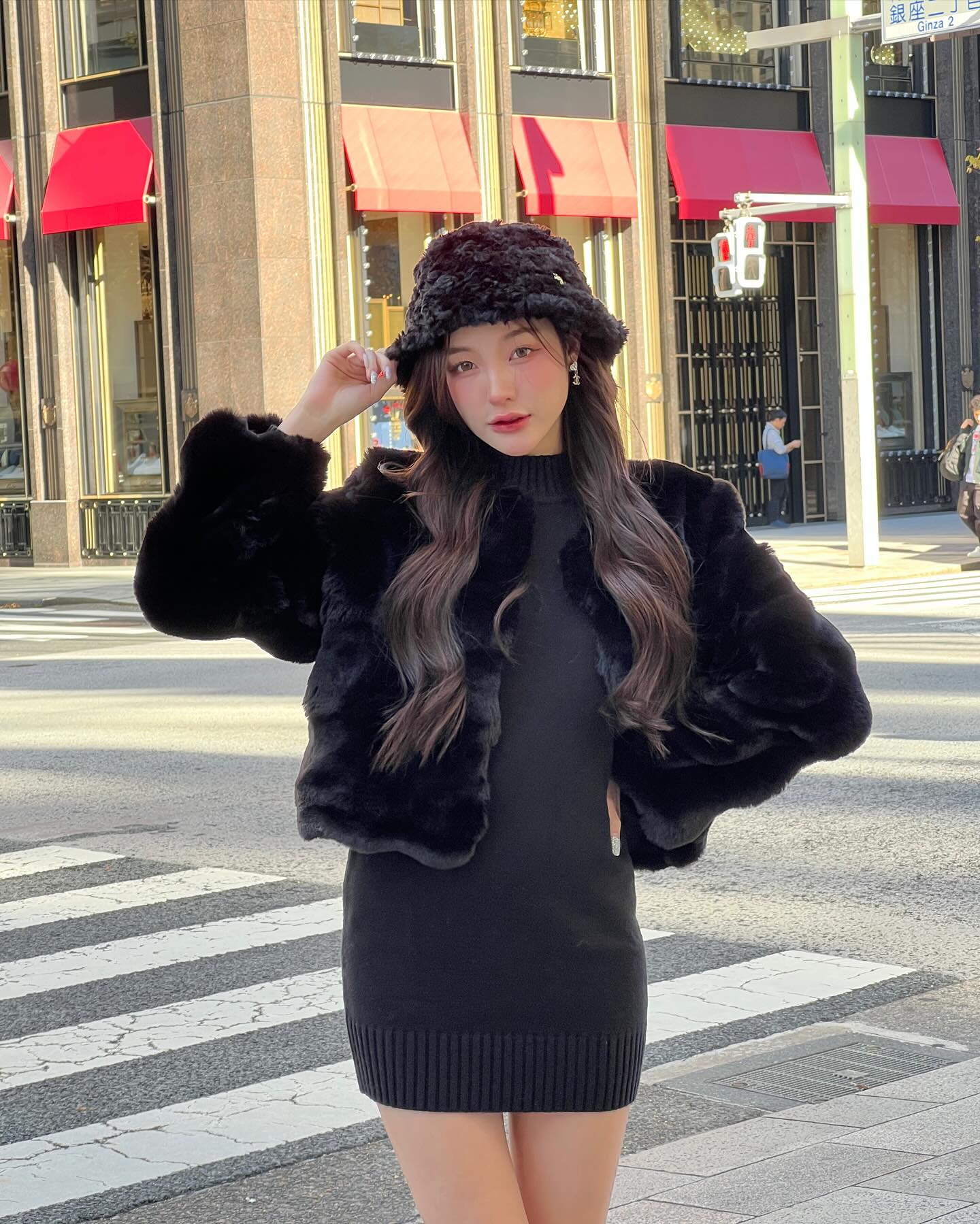 一位女士穿著 the.lookbook.select 的棉花糖毛皮大衣連帽子，站在城市的人行道上，靠近紅色遮陽篷，一身黑衣。.