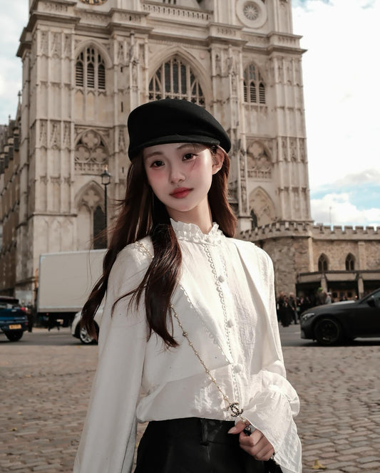 一位身穿 the.lookbook.select 的 Hazel White Shirt 🖤新品優惠至 12 月 5 日，頭戴黑色貝雷帽的女士站在鵝卵石街道上，面前是一座宏偉的哥德式大教堂。.