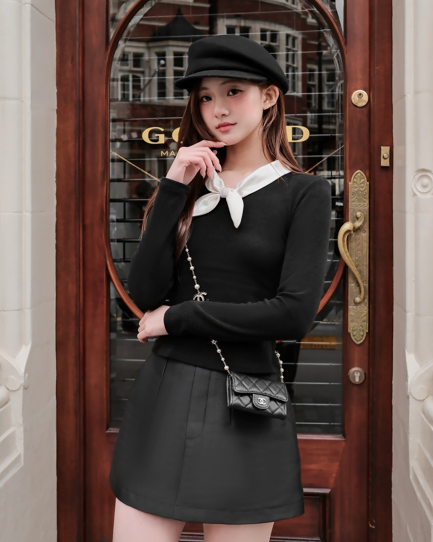 一位穿著 the.lookbook.select 的 Dear Knit Top 🖤新品優惠至 12 月 5 日、黑色短裙、貝雷帽和小挎包的女士站在一扇有玻璃面板和銅柄的木門前。.