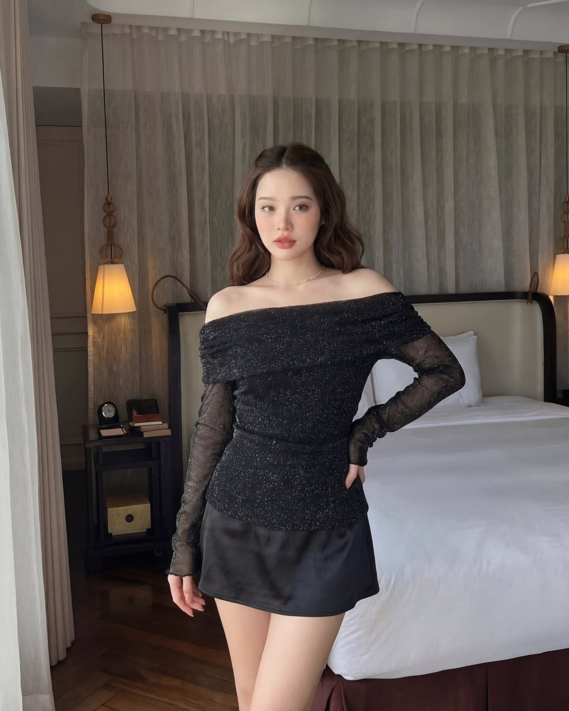 一位女士站在室內的床邊，身穿閕閕新品優惠2件9折露肩剪裁針織上衣和黑色短裙。背景懸掛兩盞吊燈。.