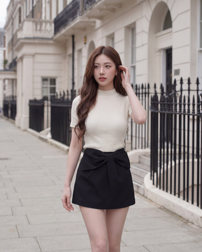 一位長髮女子身穿米色上衣，搭配 the.lookbook.select 的 Chloe 蝴蝶結半身裙，正走在城市人行道上，身後是配有黑色欄杆的白色建築。.