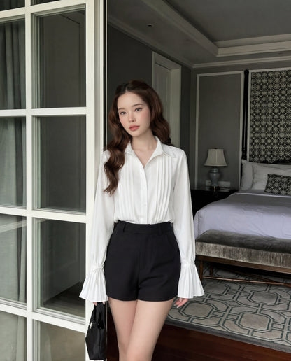 一位女士站在室內玻璃門附近，身穿 the.lookbook.select 的白色喇叭袖襯衫和黑色短褲；喇叭袖增添優雅氣質。背景是一張床和床頭櫃。.