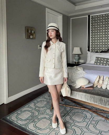 一位女士站在時尚的臥室中，身穿 the.lookbook.select 的 College Tweed Coat，搭配裙子、帽子、鞋子和包包。四對鞋子和一頂帽子整齊地排列在床邊的長椅上。.