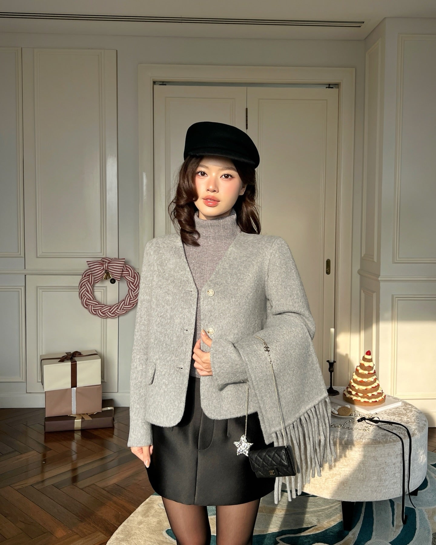 一位女士穿著.lookbook.select Snow Wool Coat❄️新品優惠和高領毛衣，搭配黑色短裙和帽子，站在室內的桌子旁，背景是蛋糕和包好的禮物。.