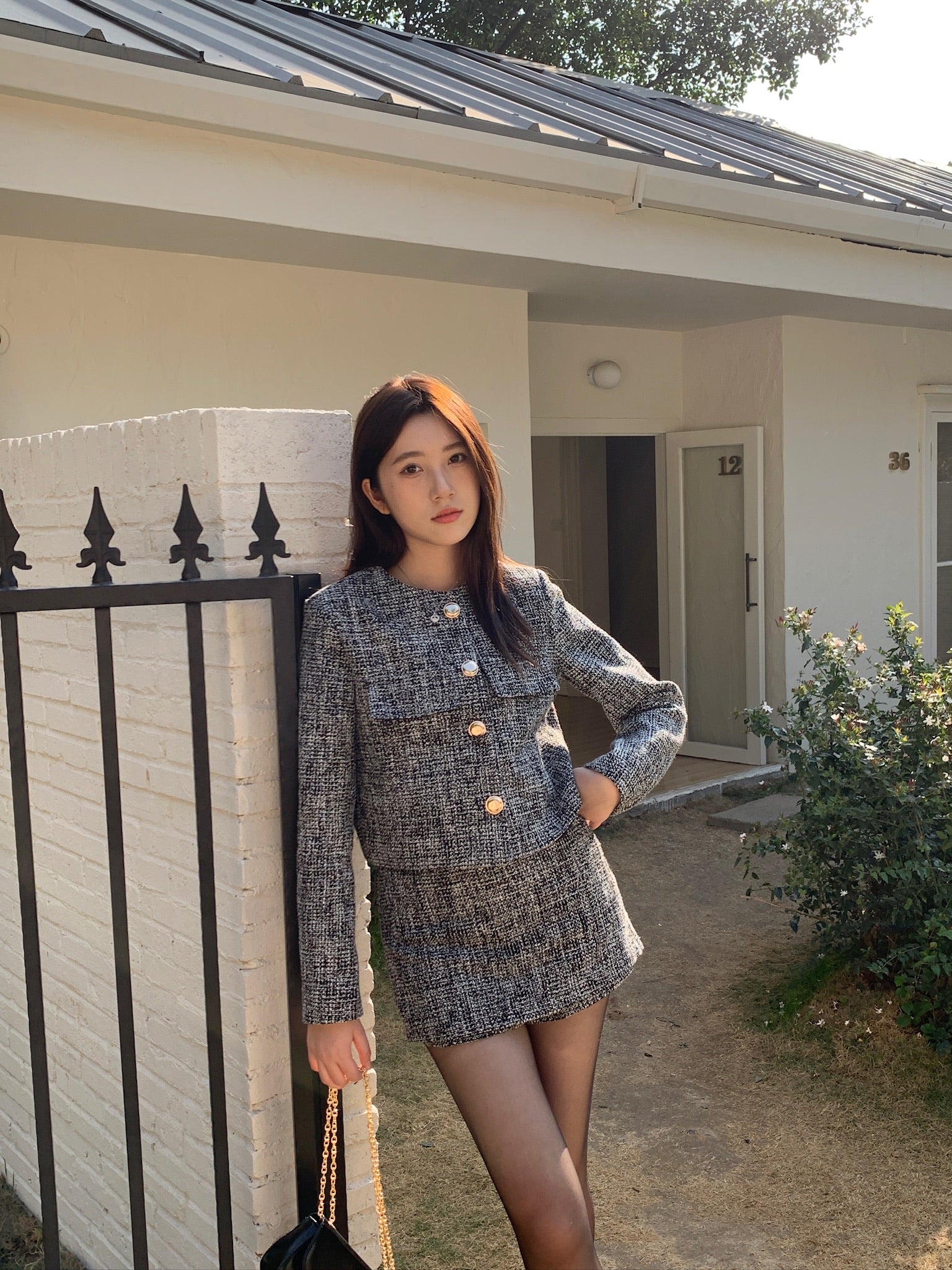 一位身穿 the.lookbook.select 的「This Girl Tweed Set」的女子站在一棟現代化房屋前，旁邊是一根帶有黑色柵欄的白磚柱子。.