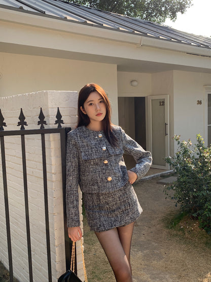 一位身穿 the.lookbook.select 的「This Girl Tweed Set」的女子站在一棟現代化房屋前，旁邊是一根帶有黑色柵欄的白磚柱子。.