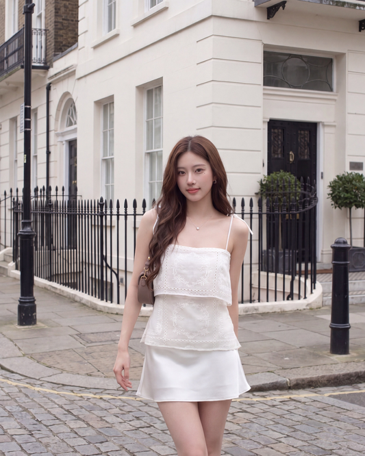 一位女士穿著the.lookbook.select的Heirloom Eyelet Cami，站在鵝卵石街道上一棟有黑色欄杆的白色建築物前。閕新品優惠2件9折.