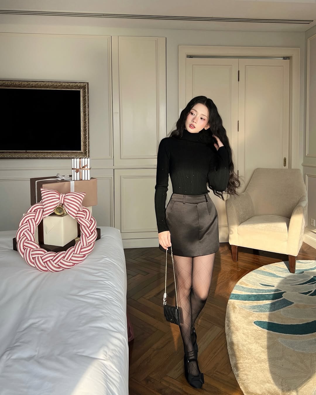 一位身穿 the.lookbook.select's Twinkle Knit Top❄️新品優惠、短裙和透明緊身褲的女士站在陽光普照的房間裡，床邊有粉紅色和白色的裝飾花圈。.