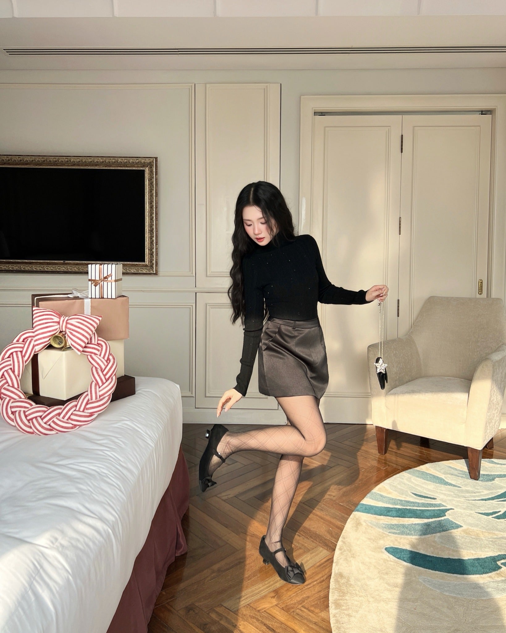 在明亮的現代房間中，一位女士身穿 the.lookbook.select 的 Twinkle Knit Top❄️，在床邊的木地板上擺姿勢，床頭擺放著禮物和花圈。.