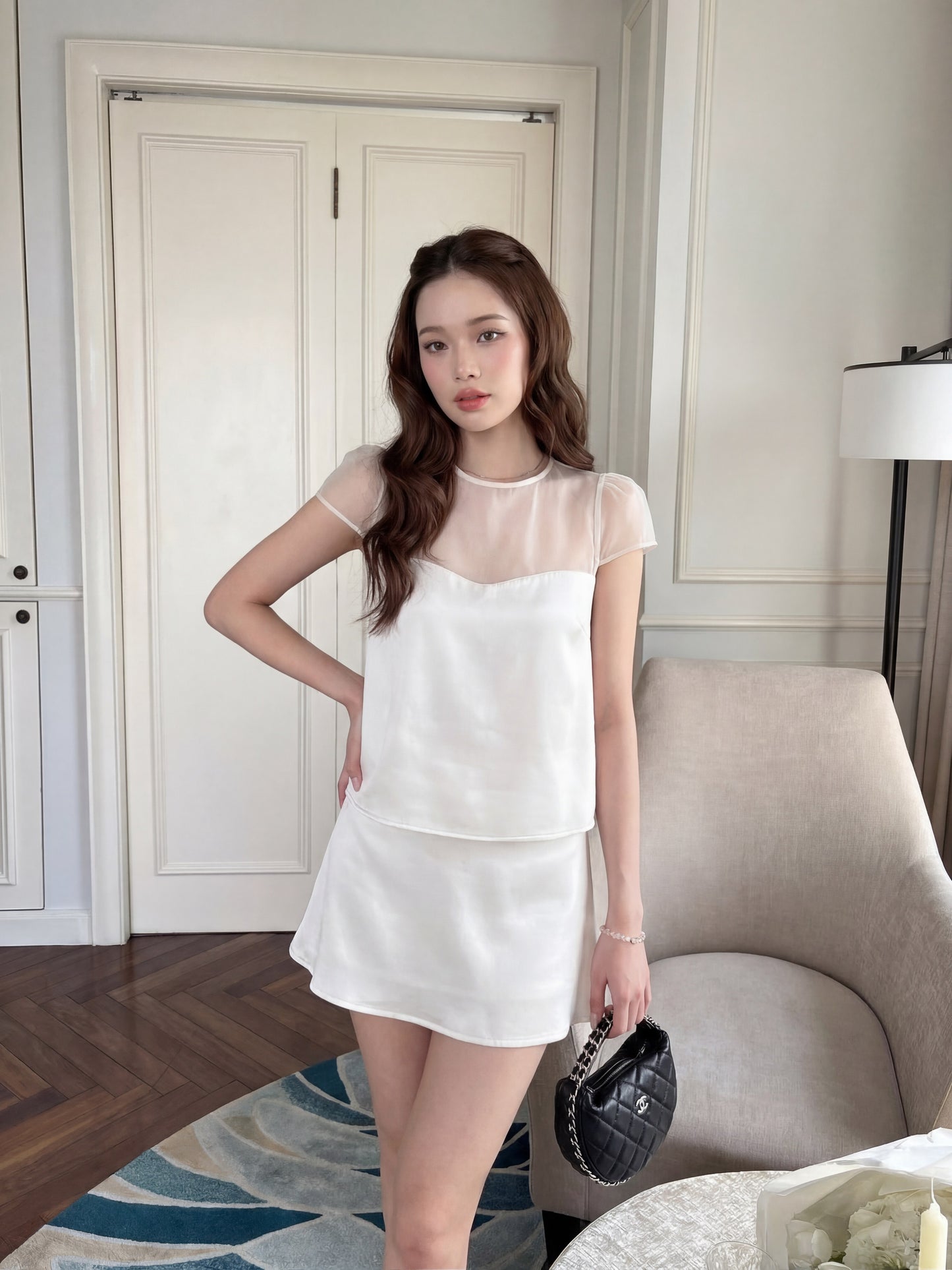 一名女子身穿 the.lookbook.select 品牌的「Satin Reverie 套裝」（上衣與裙子），手拿黑色手提包，一隻手搭在臀部，站在米色扶手椅和落地燈旁。.