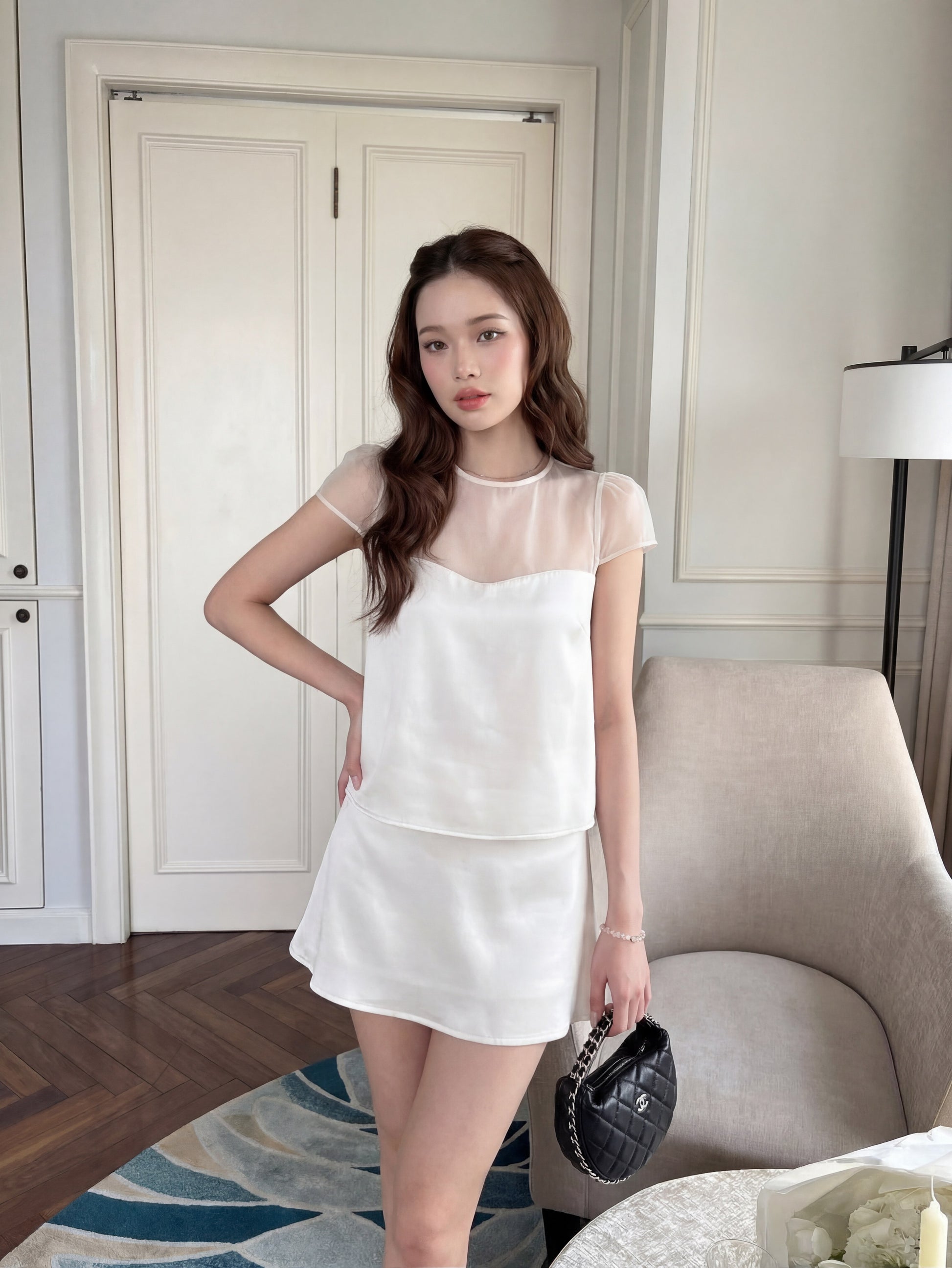 一名女子身穿 the.lookbook.select 品牌的「Satin Reverie 套裝」（上衣與裙子），手拿黑色手提包，一隻手搭在臀部，站在米色扶手椅和落地燈旁。.