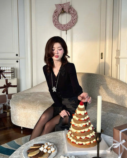 一位身穿 the.lookbook.select 蕾絲針織上衣的女士坐在米色沙發上，伸手去拿桌上的層次分明的聖誕樹形蛋糕，旁邊是餅乾和包好的禮物。.