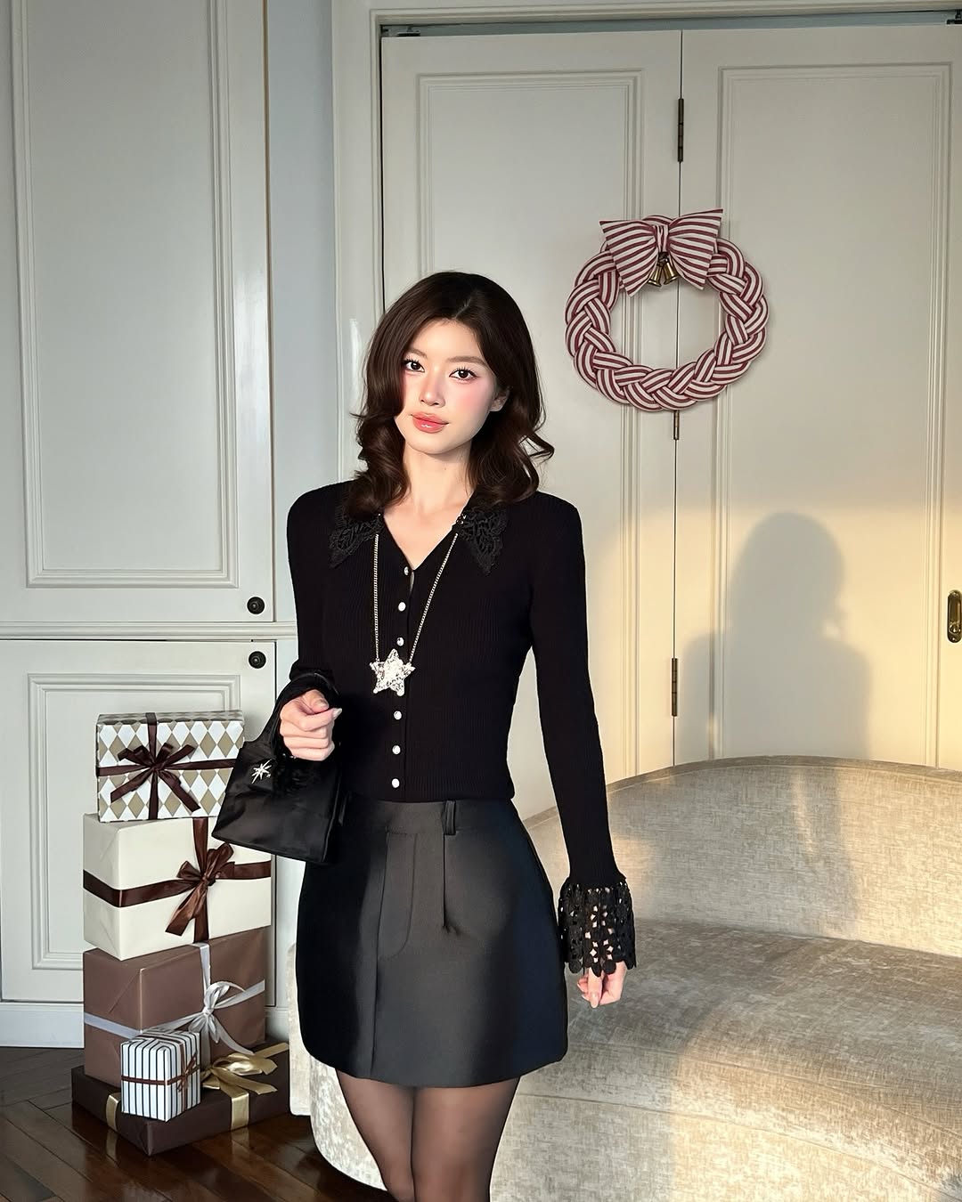 一位女士在室內沙發旁穿著 the.lookbook.select 的蕾絲針織上衣，附近堆疊著包裝好的禮物，背後的門上掛著紅白相間的花圈。.