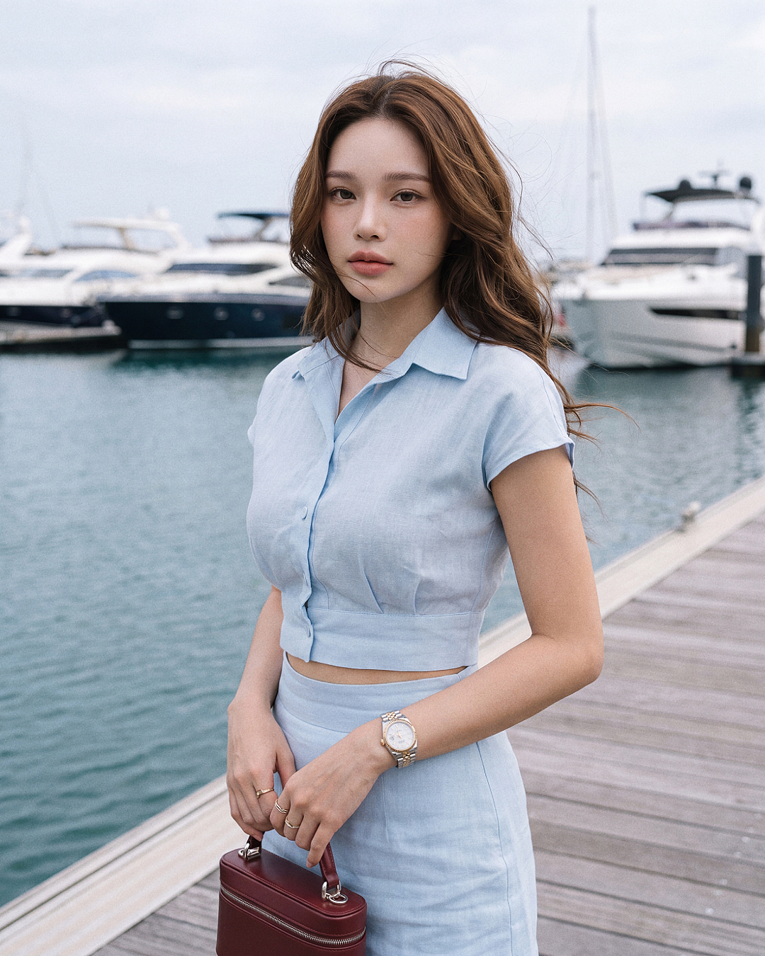 一位身穿 the.lookbook.select「Breeze 亞麻套裝」的女子站在碼頭上，身後是遊艇與水面，手中提著一個小巧的栗色手提包。.