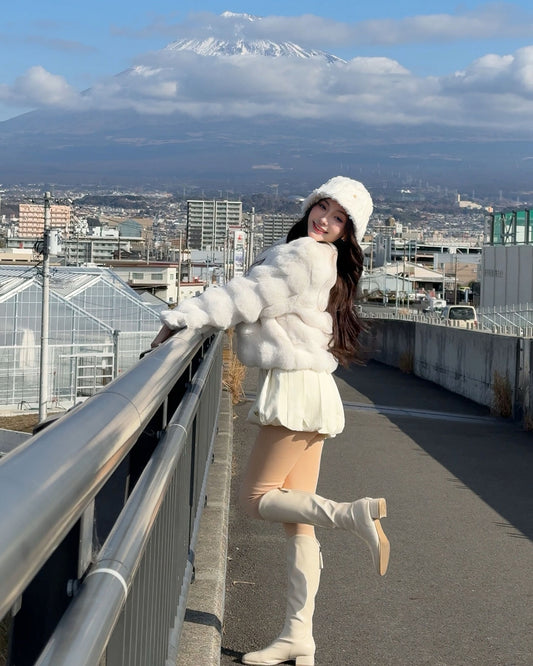 一位女士在橋上為 the.lookbook.select 的棉花糖皮草大衣 (附帽子) 當模特兒，背景是部分陰天下的城市建築和雪山。.