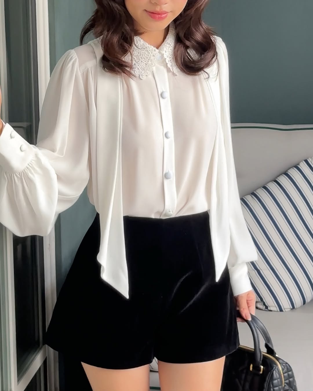 一位女士身穿.lookbook.select Heart White Shirt ❄️ 新品優惠，蕾絲領口搭配黑色高腰短褲，手拿黑色手提包，站在室內有條紋靠墊的沙發旁。.