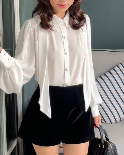 一位女士身穿.lookbook.select Heart White Shirt ❄️ 新品優惠，蕾絲領口搭配黑色高腰短褲，手拿黑色手提包，站在室內有條紋靠墊的沙發旁。.