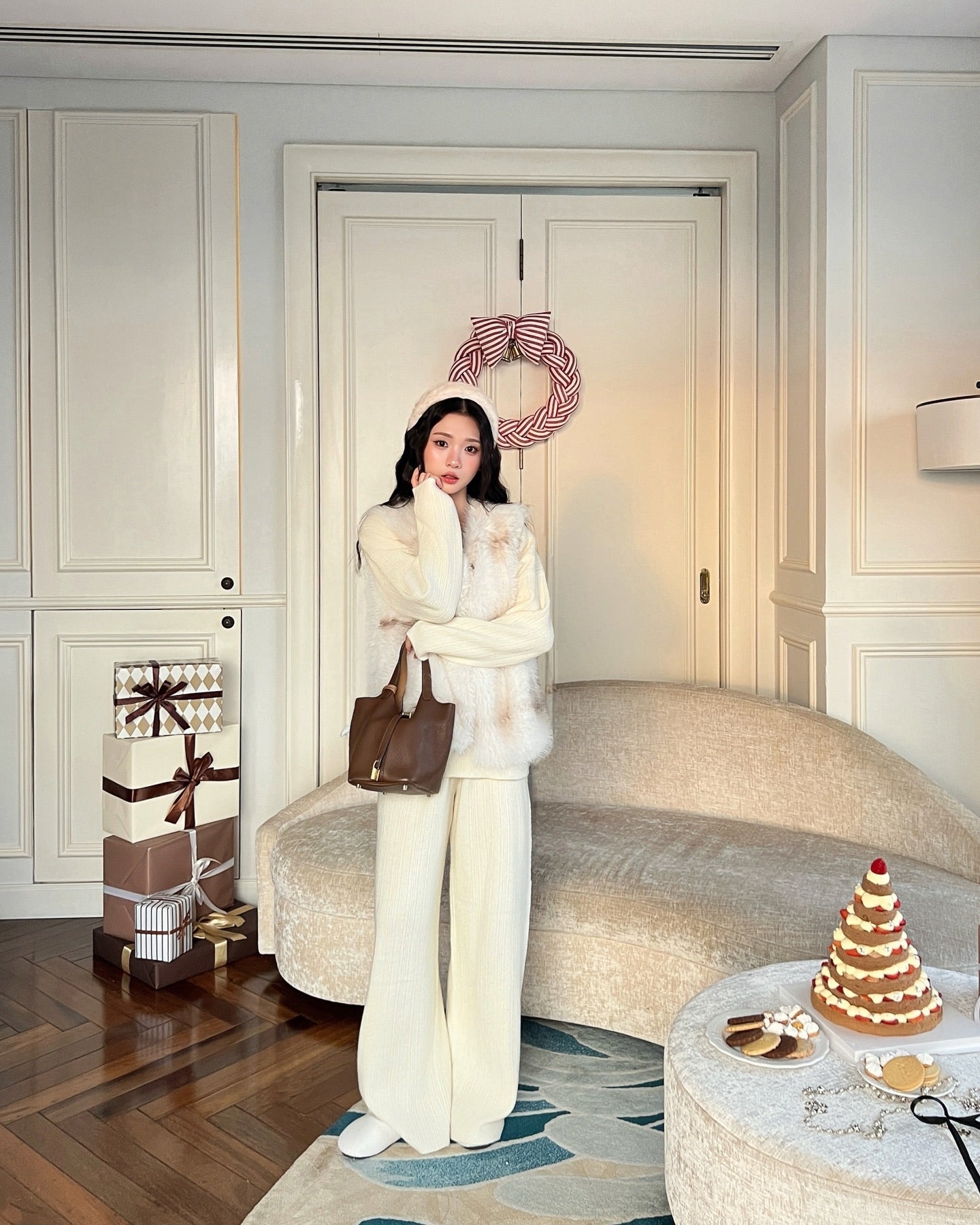 一位身著乳白色冬季服飾的女士站在裝潢充滿節日氣氛的客廳中，她身後的門上掛著禮物、甜點和花圈，她穿著由 the.lookbook.select 製作的 La Neige Fur Vest ❄️ 新品優惠。.
