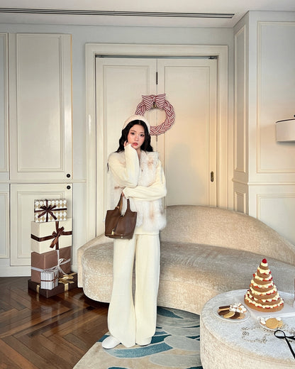 一位身著乳白色冬季服飾的女士站在裝潢充滿節日氣氛的客廳中，她身後的門上掛著禮物、甜點和花圈，她穿著由 the.lookbook.select 製作的 La Neige Fur Vest ❄️ 新品優惠。.