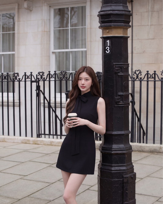 一位身穿 the.lookbook.select 的 Estate 領結迷你裙的女士站在城市人行道上的黑色燈柱旁，手持咖啡杯，背景是鐵欄桿和建築物。.