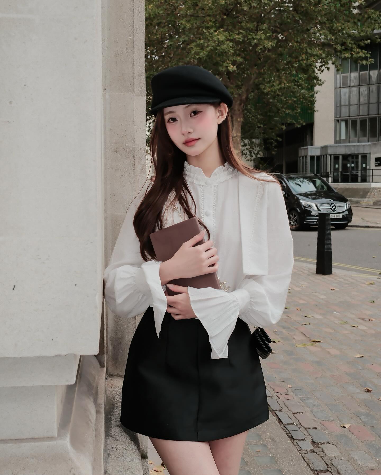 一位身穿白色上衣、the.lookbook.select Grace Mini Skirt 🖤新品優惠至 12 月 5 日、頭戴黑色貝雷帽的女士倚靠著石牆，手持棕色手拿包走在城市的人行道上。.